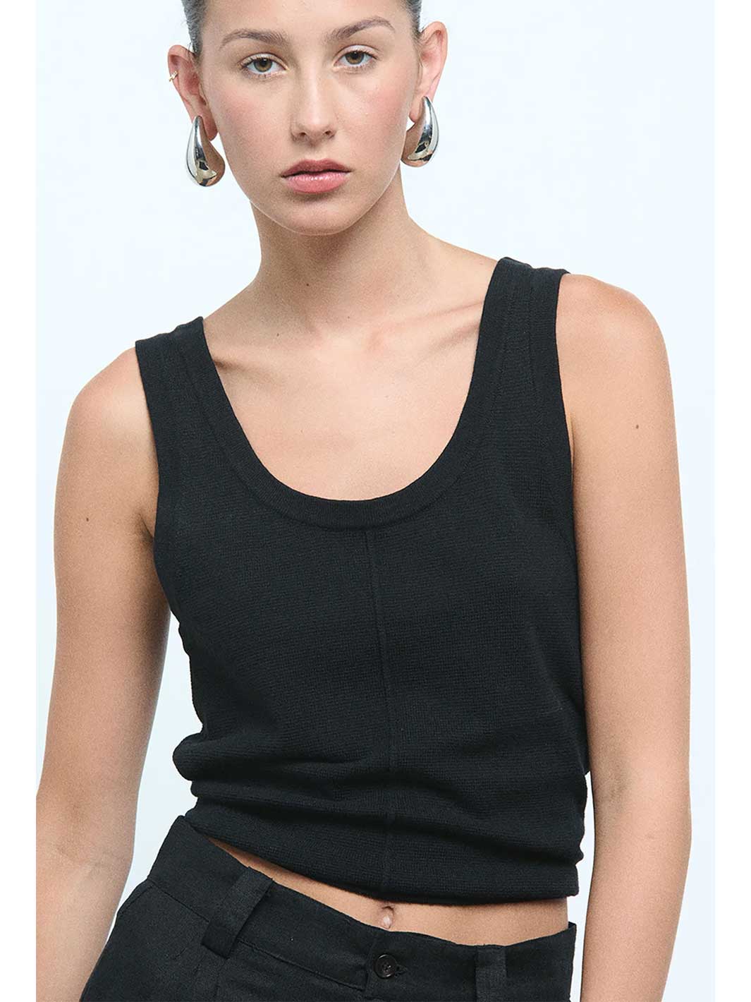 A woman wearing a black ribbed knit scoop-neck tank top and black pants, styled with large silver hoop earrings against a light blue studio background.