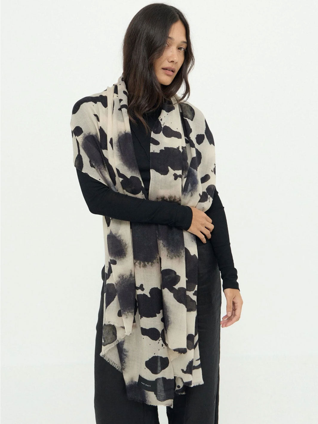 A woman modeling a long, lightweight wrap or shawl with a black and off-white cow-print pattern, worn over a black long-sleeve top and black pants against a plain white studio background.