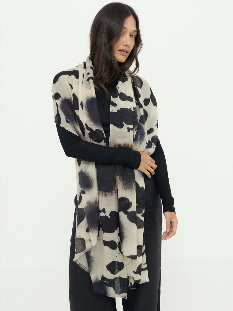A woman modeling a long, lightweight wrap or shawl with a black and off-white cow-print pattern, worn over a black long-sleeve top and black pants against a plain white studio background.