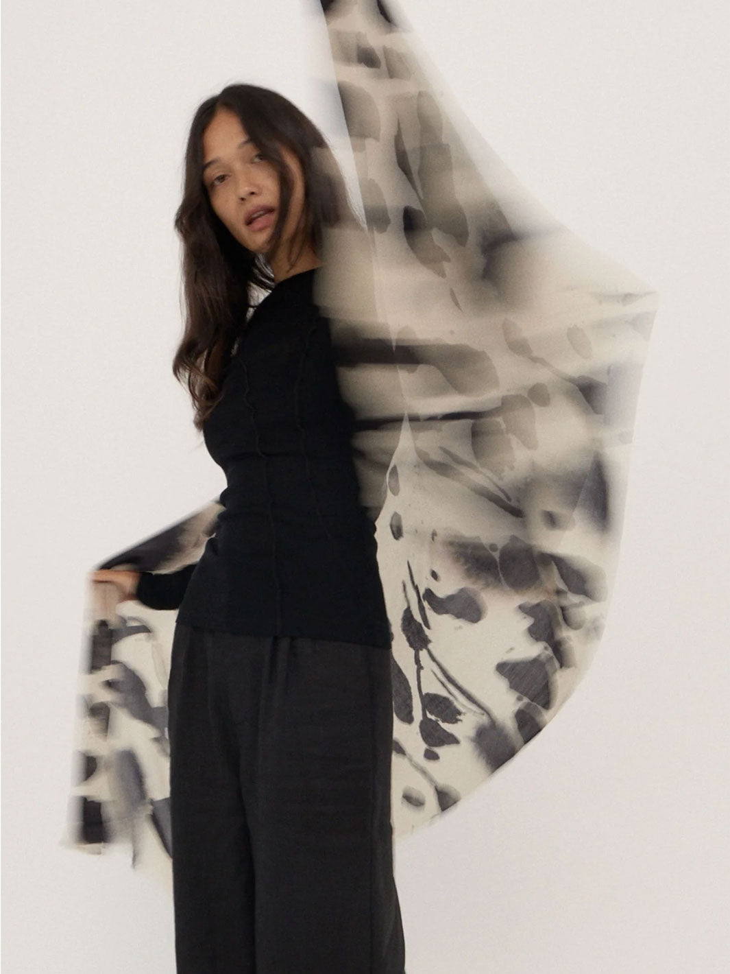 A woman modeling a sheer black and white abstract patterned scarf or shawl draped over her shoulders, paired with a black top and dark wide-leg pants against a plain white studio background.