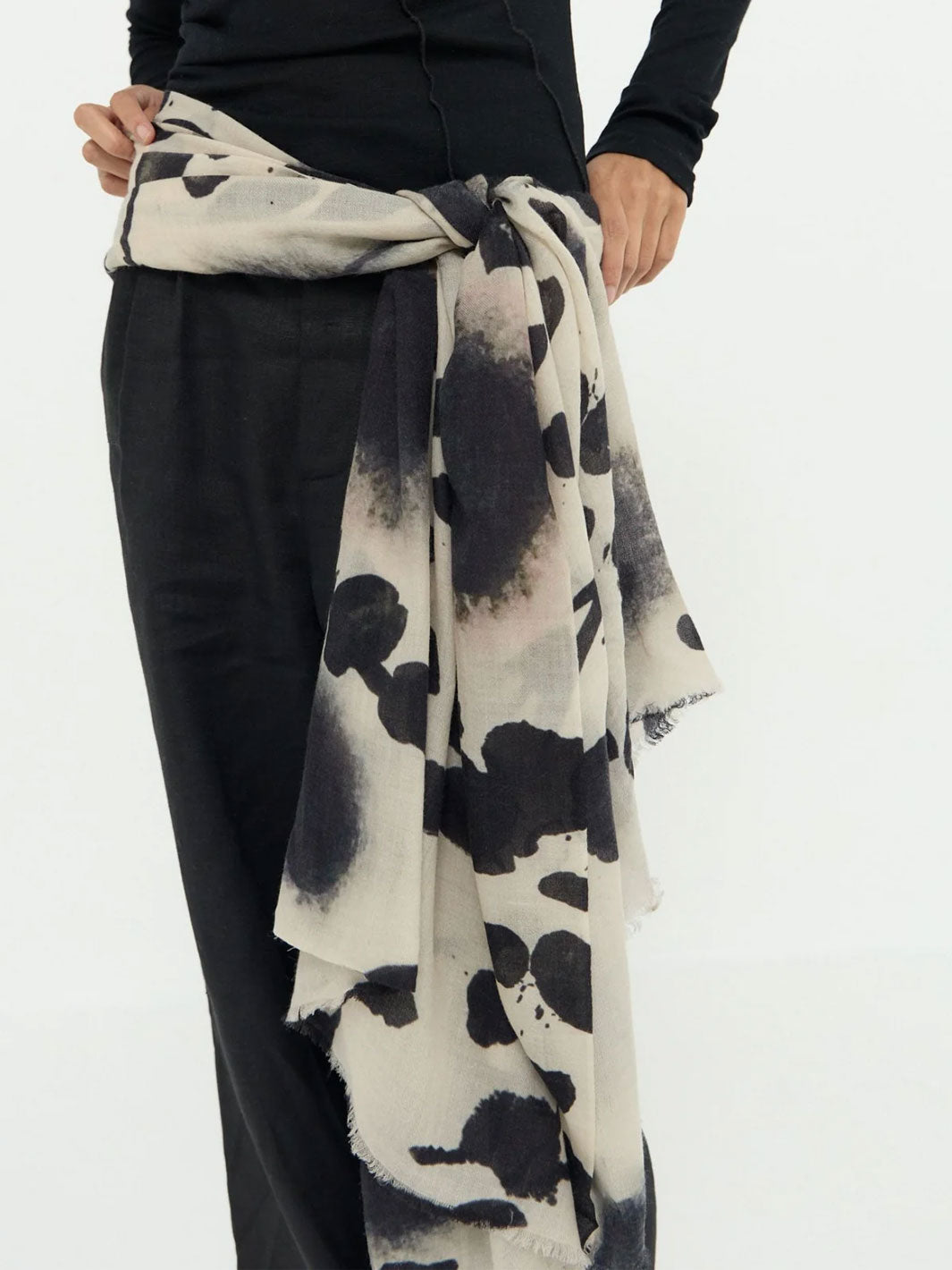 A person wearing a black long-sleeve top and black pants, shown from the chest down, with a large lightweight scarf tied around the waist. The scarf has an abstract black and off-white watercolor or ink-blot pattern.