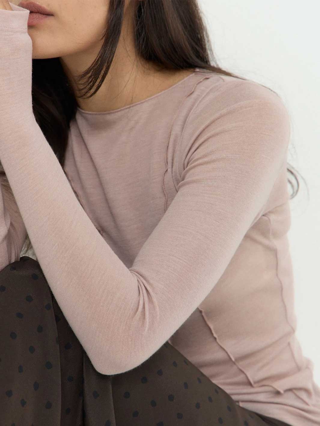 A close-up of a woman wearing a fitted, long-sleeve crew-neck top in a soft dusty pink or mauve shade, paired with a dark brown polka-dot skirt against a plain white background.