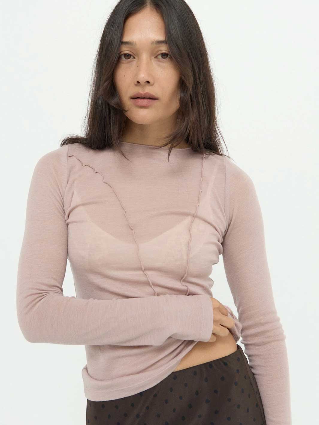A woman modeling a long-sleeve ribbed knit top in a dusty mauve-pink color, styled with a sheer underlayer and a dark patterned skirt against a plain white background.