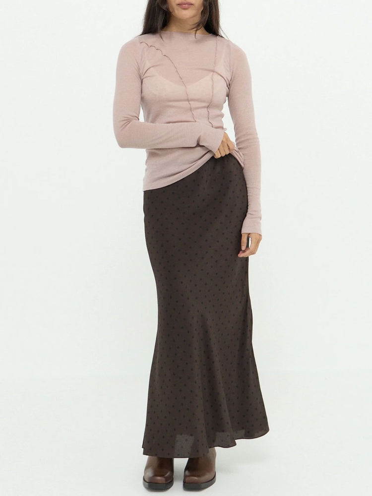 A model wearing a light pink mock-neck knit top and a dark brown skirt with a small polka-dot pattern, standing against a plain white background.