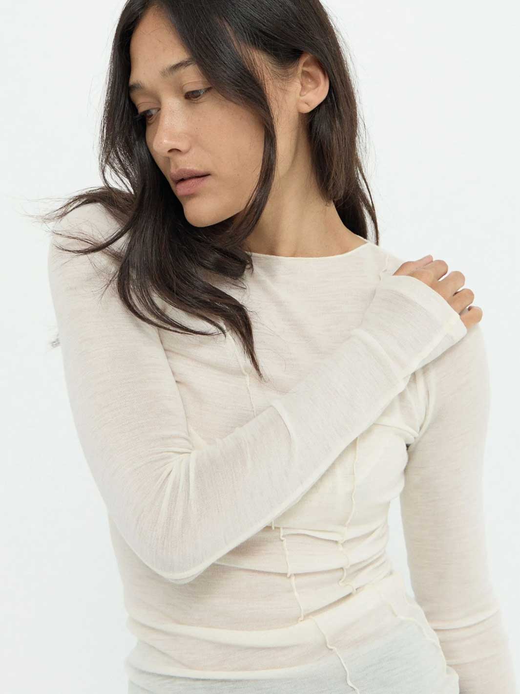 A woman wearing a fitted, long-sleeve cream-colored knit top with asymmetric seam detailing at the waist, photographed against a plain white studio background.