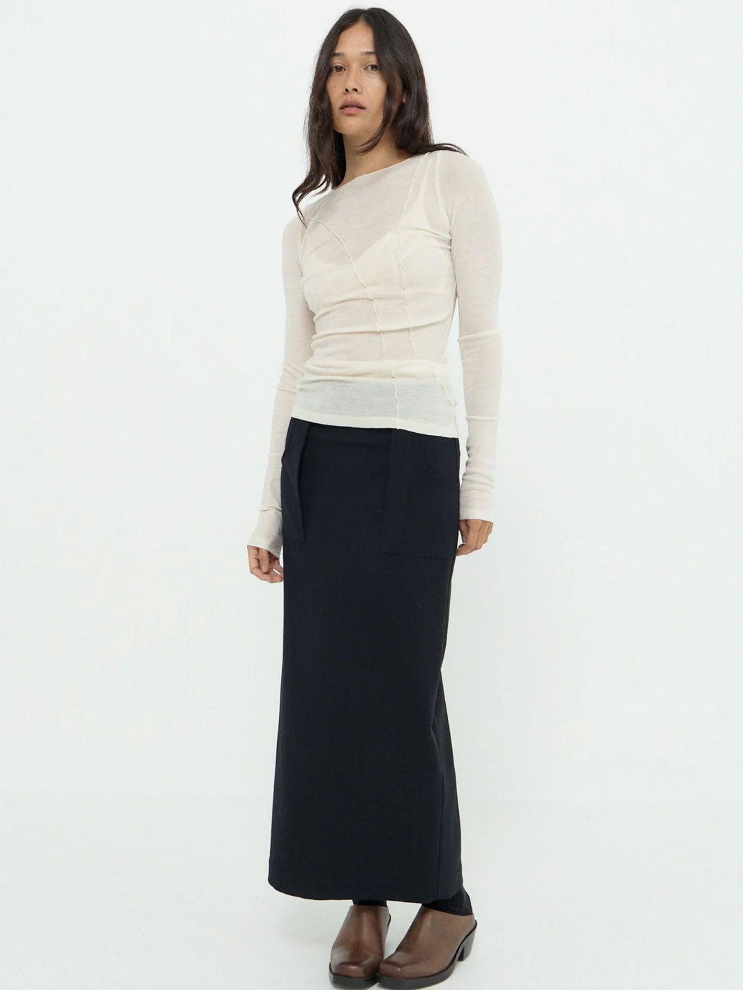 A woman modeling a cream-colored long-sleeve knit top with a sheer overlay and a black maxi skirt, paired with brown leather shoes, against a plain white studio background.