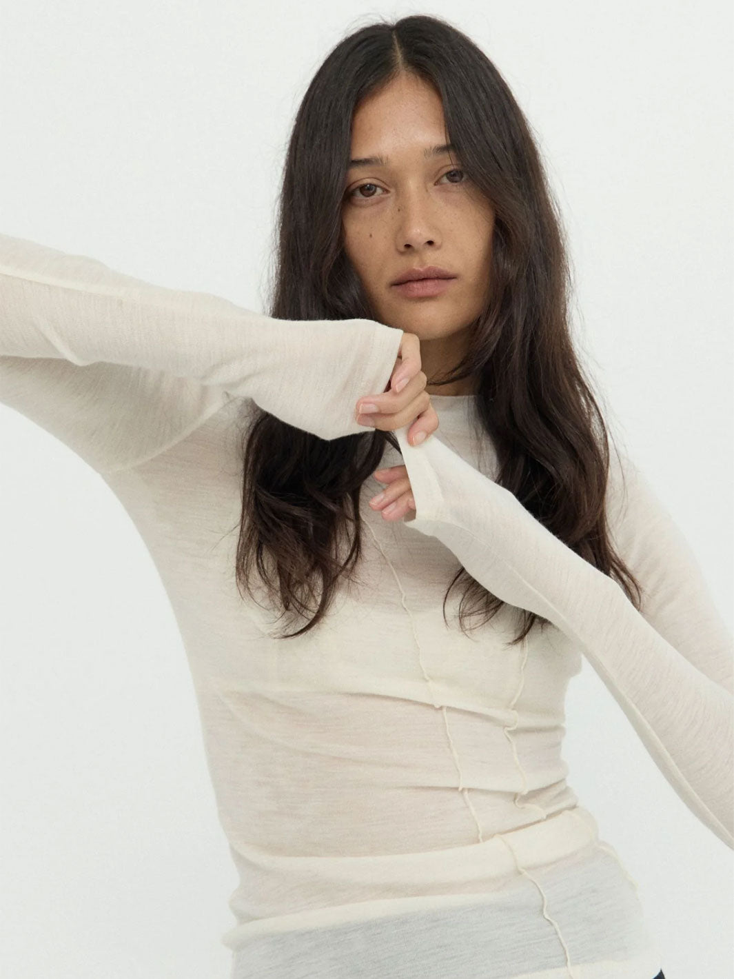 A woman wearing a fitted, long-sleeve cream-colored knit top with seamdetailing at the waist, posed against a plain white studio background.