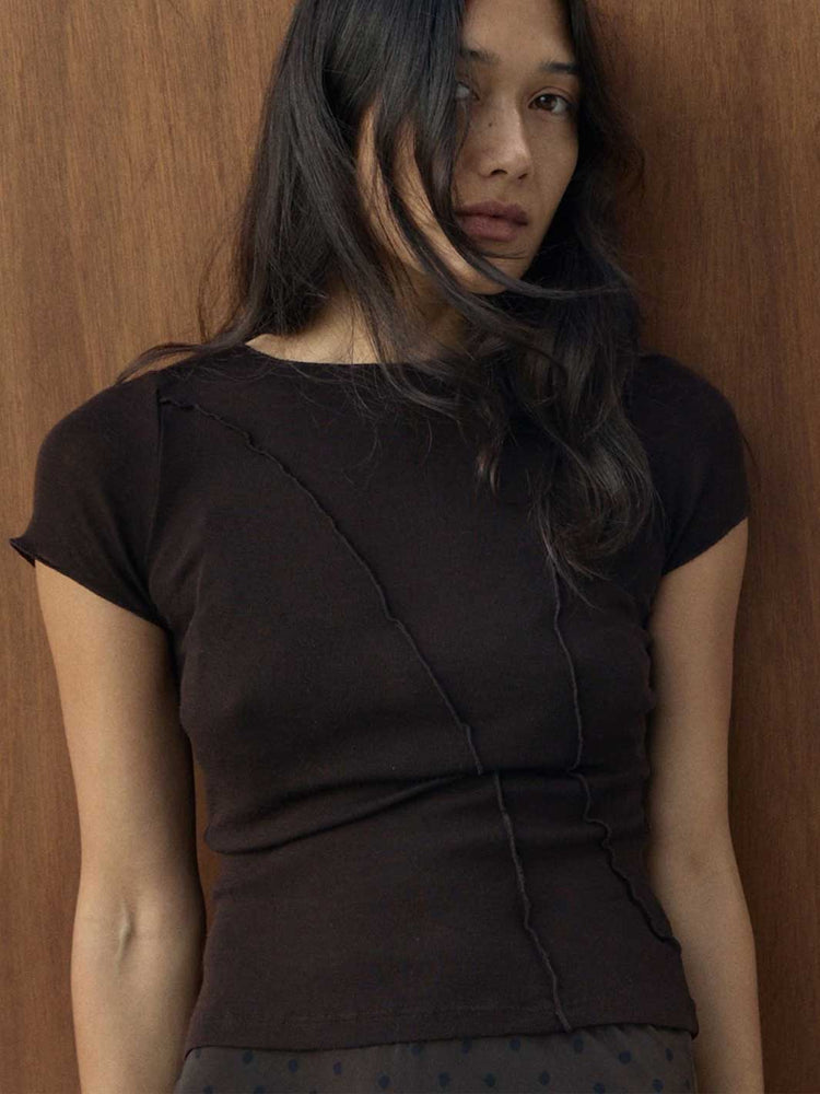 A woman wearing a dark brown short-sleeve top with diagonal seam details, leaning against a wooden wall.
