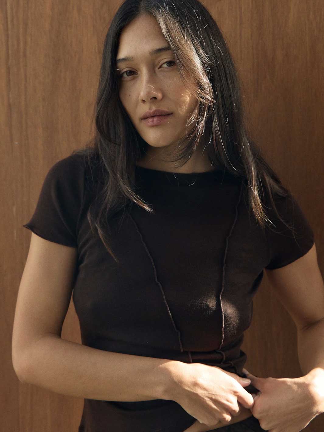 A woman with long dark hair wearing a tight dark brown short-sleeve top, standing against a warm brown wooden wall.