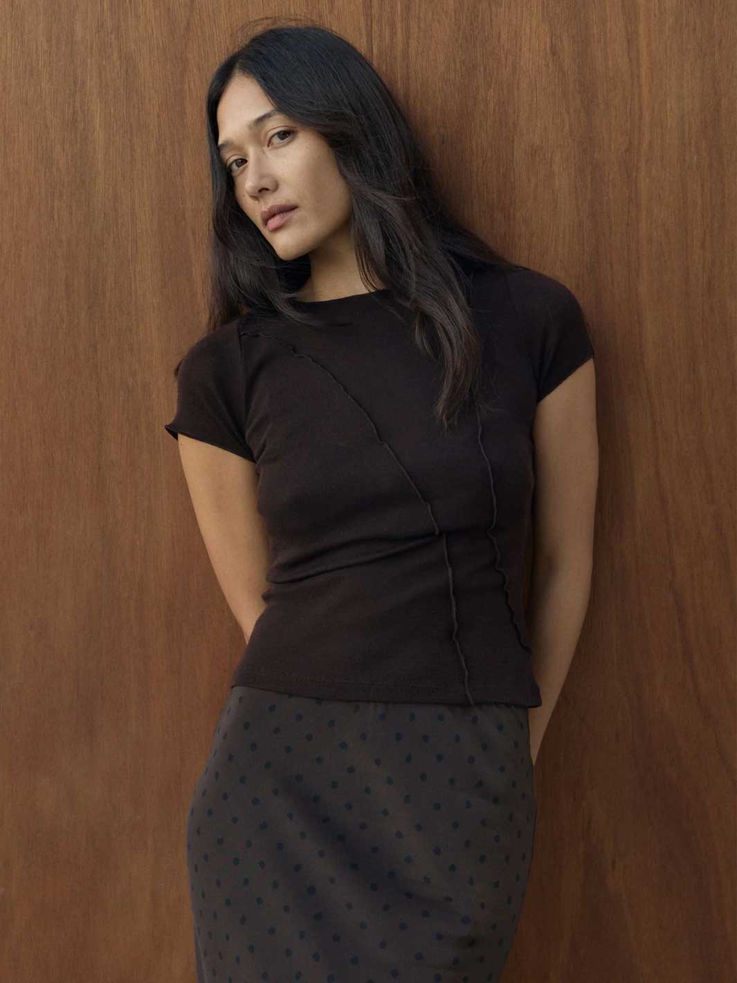 A woman with long dark hair leans against a brown wooden wall, wearing a fitted dark brown short-sleeve top and a dark grey skirt with small black polka dots.