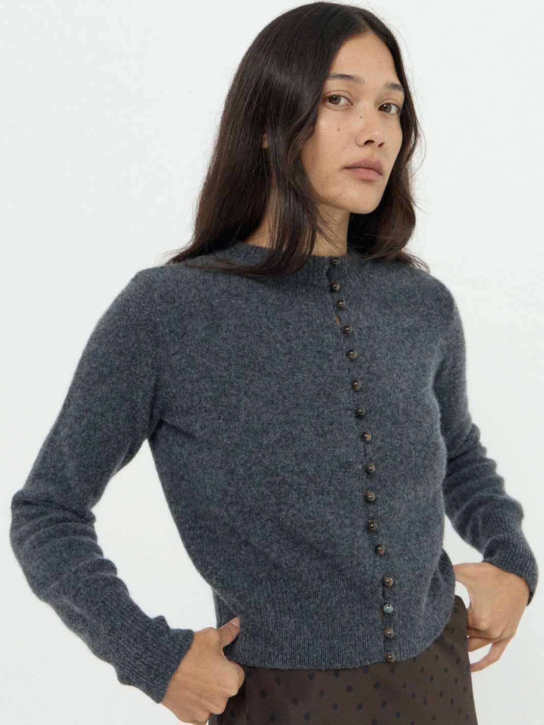 A woman wearing a dark gray knit button-front cardigan sweater, paired with a dark patterned skirt, against a plain white studio background.