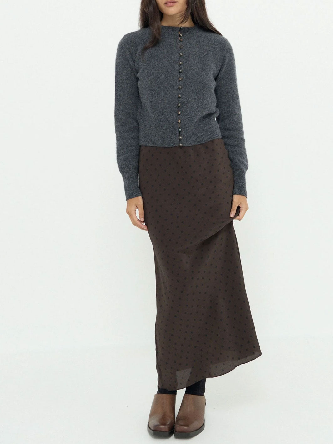 A model wearing a dark gray knit button-front cardigan and a dark brown midi skirt with a small polka-dot pattern, paired with brown leather boots, against a plain white studio background.