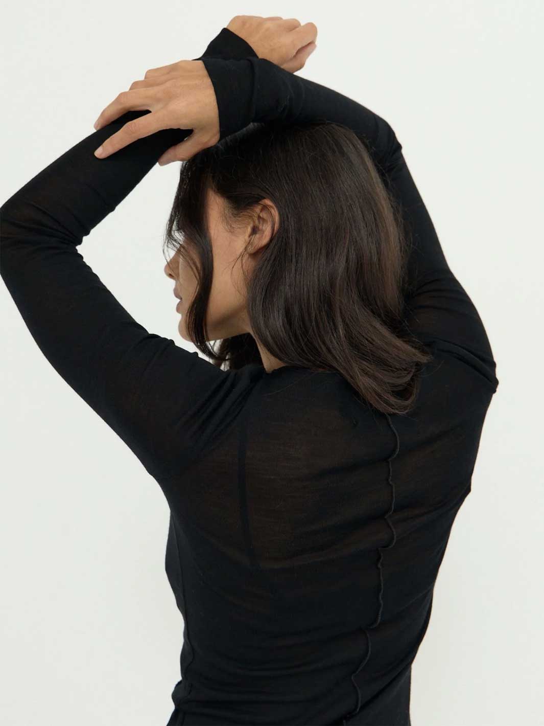 A woman wearing a black long-sleeve sheer merino knit top, shown from the back as she stretches her arms overhead, against a plain white background.