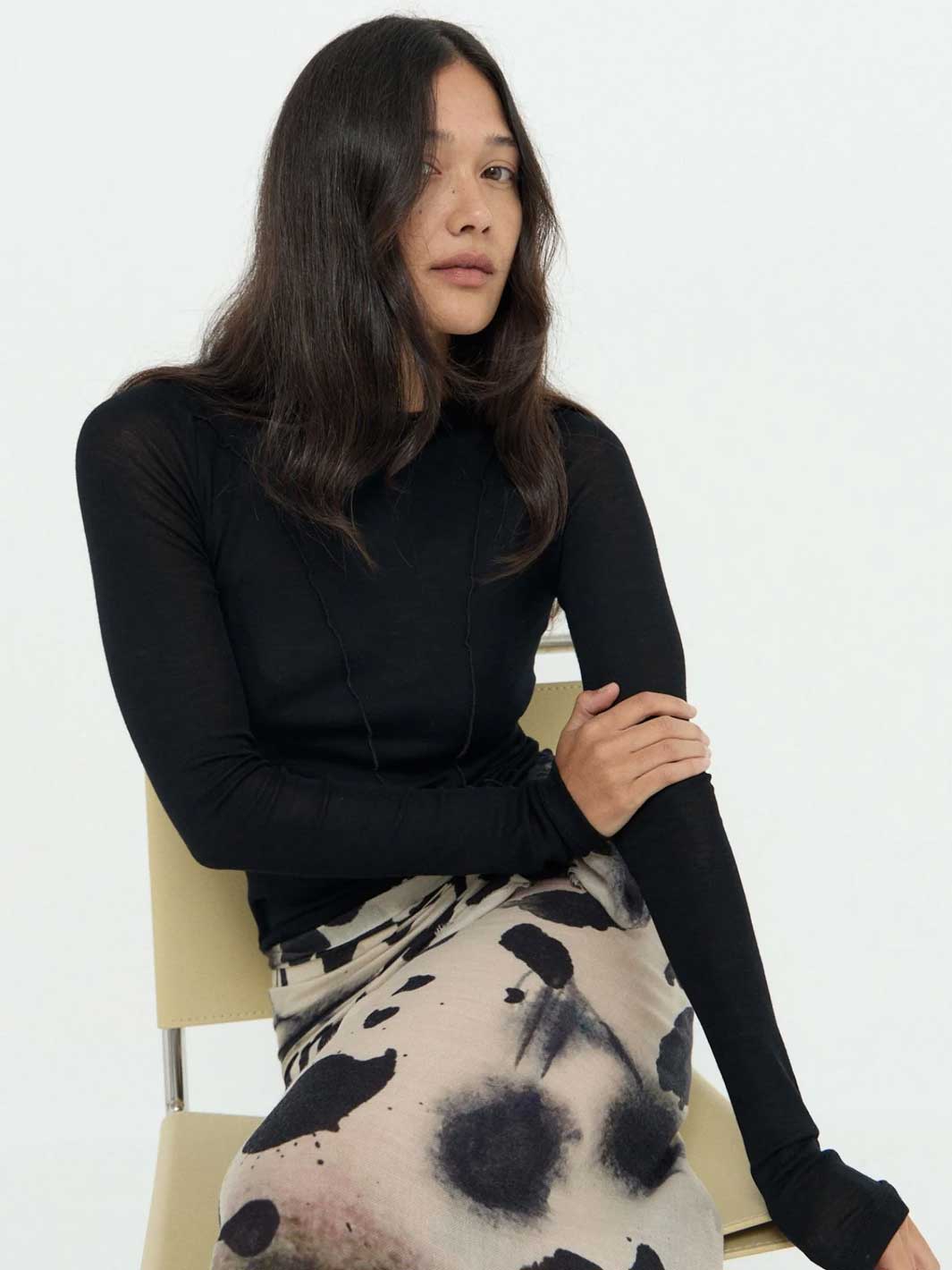 A woman seated on a light beige chair wearing a fitted black long-sleeve top and a beige skirt with a black abstract print, photographed against a plain light background.