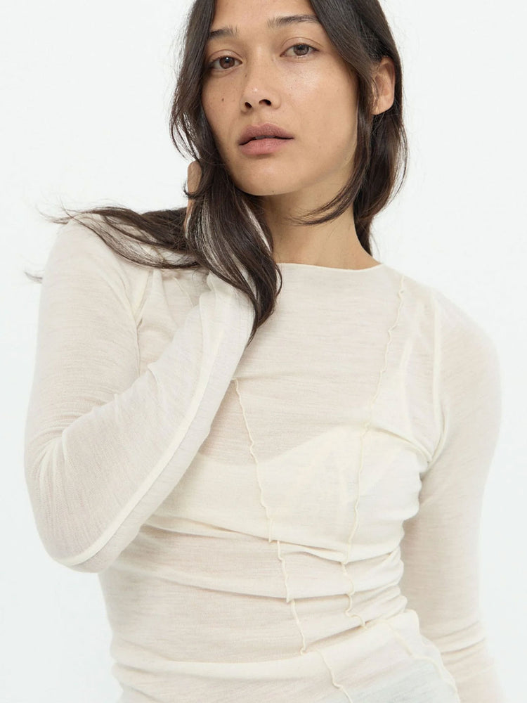 A woman wearing a fitted, long-sleeve cream-colored top with gathered details and a boat neckline, photographed against a plain white background.