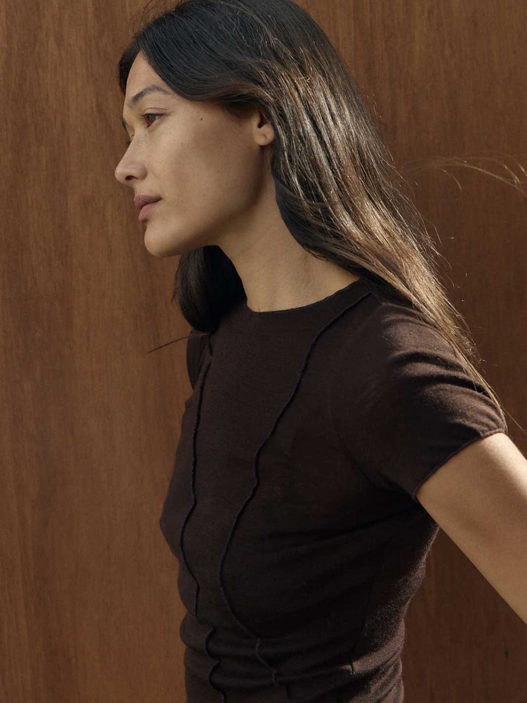 A woman in profile wearing a dark brown short-sleeve knit top with vertical seam details, set against a warm brown wood-paneled background.