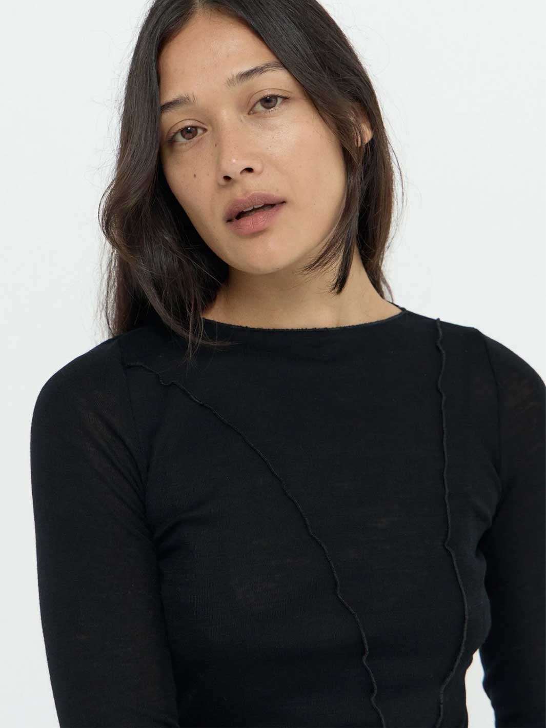 A woman wearing a black long-sleeve knit top with a boat neckline and subtle vertical seam details, photographed against a plain light background.