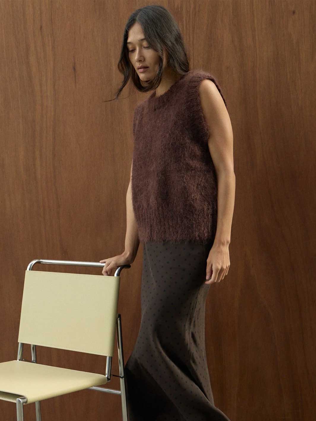 A woman stands indoors against a wood-paneled wall, wearing a sleeveless fuzzy brown top and a dark skirt. She is holding the back of a cream-colored chair with a metal frame.