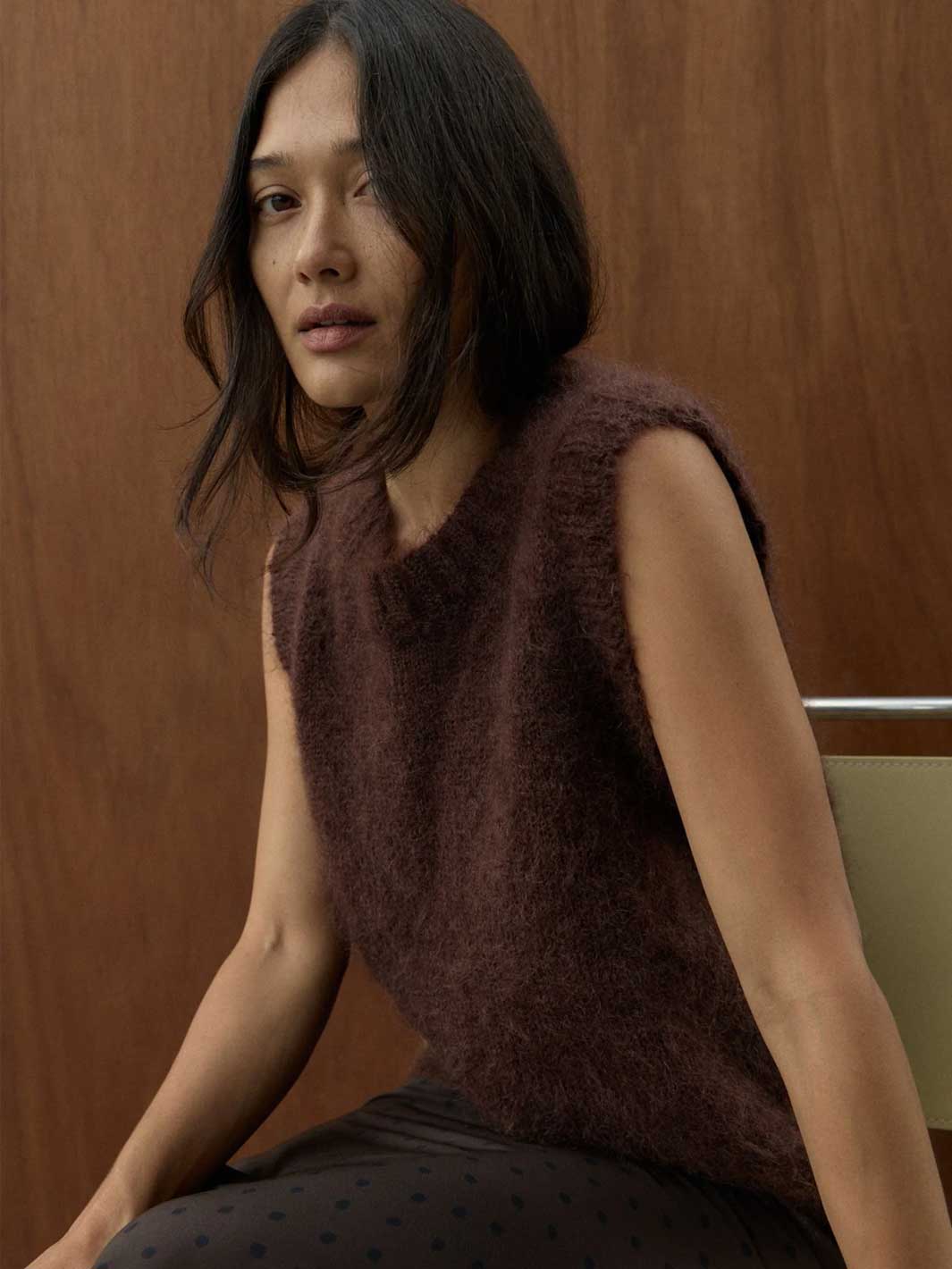 A woman wearing a sleeveless, fuzzy brown knit top and dark patterned pants, seated against a warm wood-paneled background.
