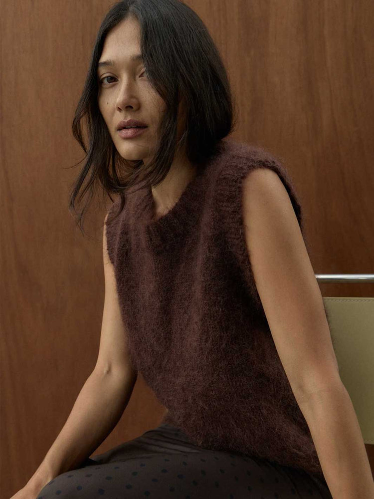 A woman wearing a sleeveless, fuzzy brown knit top and dark patterned pants, seated against a warm wood-paneled background.