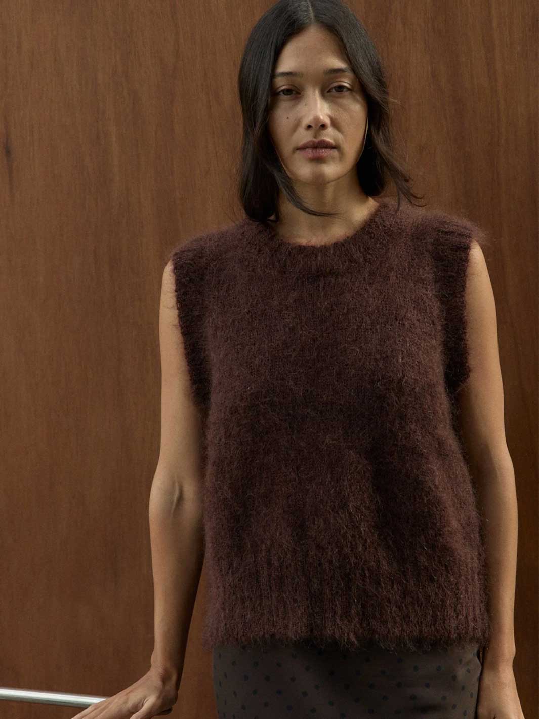 A woman wearing a sleeveless, fuzzy brown knit top with a crew neck, standing against a wooden wall.