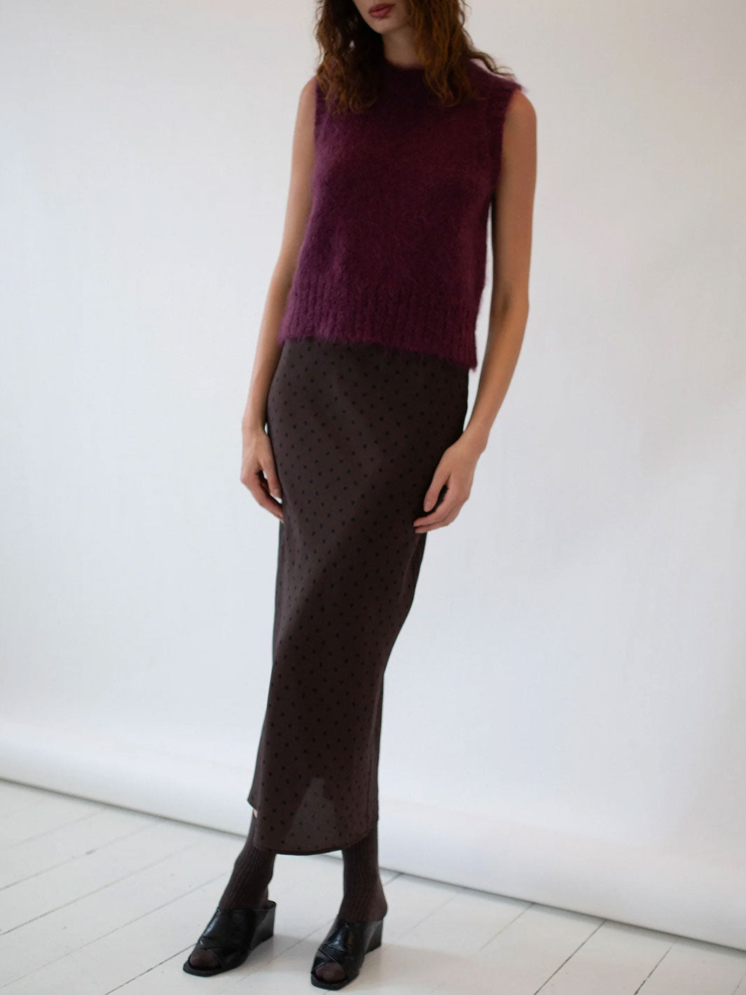 Person wearing a dark purple sleeveless top and brown patterned skirt against a white background