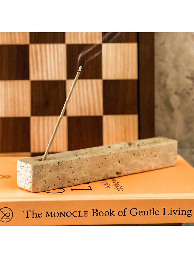 A rectangular stone incense holder with a smoking incense stick inserted, displayed on top of an orange book with the title 'The Monocle Book of Gentle Living' visible. A wooden checkerboard and textured wall appear in the background.