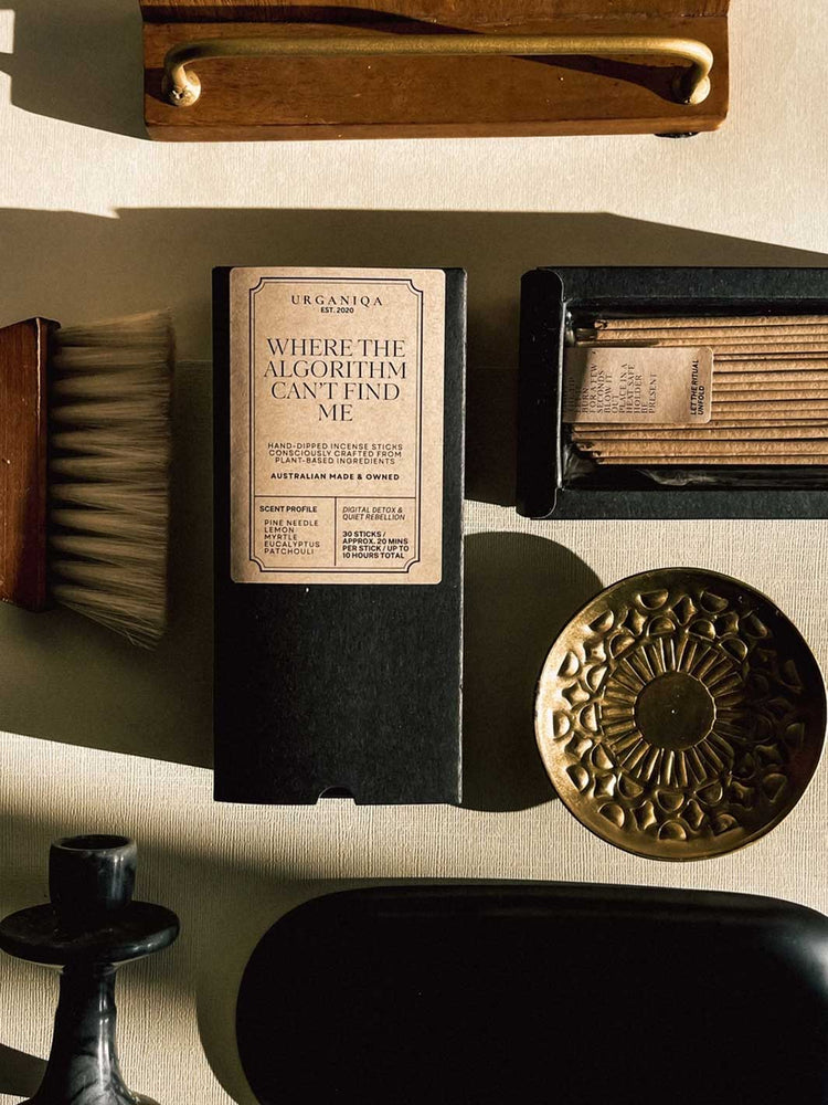 A styled flat-lay of a black incense box with a beige label reading 'Where the Algorithm Can't Find Me' by Uganiqa, surrounded by grooming props on a light textured surface.