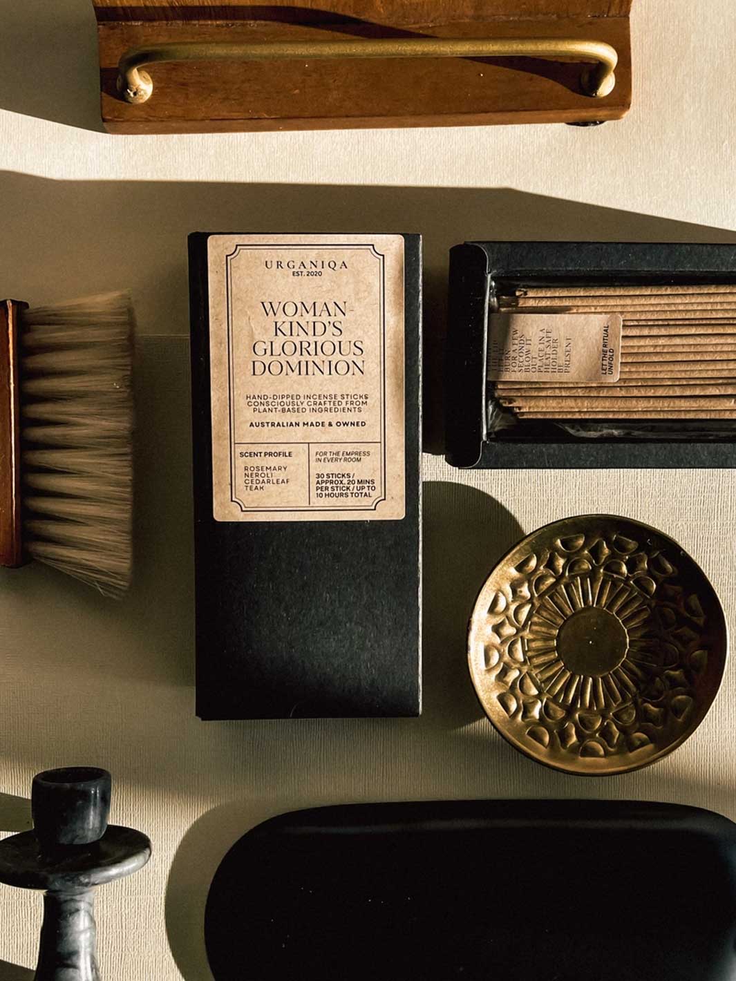 A styled flat-lay featuring a black rectangular box with a cream label that reads 'URGANIQA THE WOMAN KIND'S GLORIOUS DOMINION' Surrounding props include a wooden handle, a brush, a tray of wooden sticks, a round brass decorative piece, and a dark oval object on a beige surface.