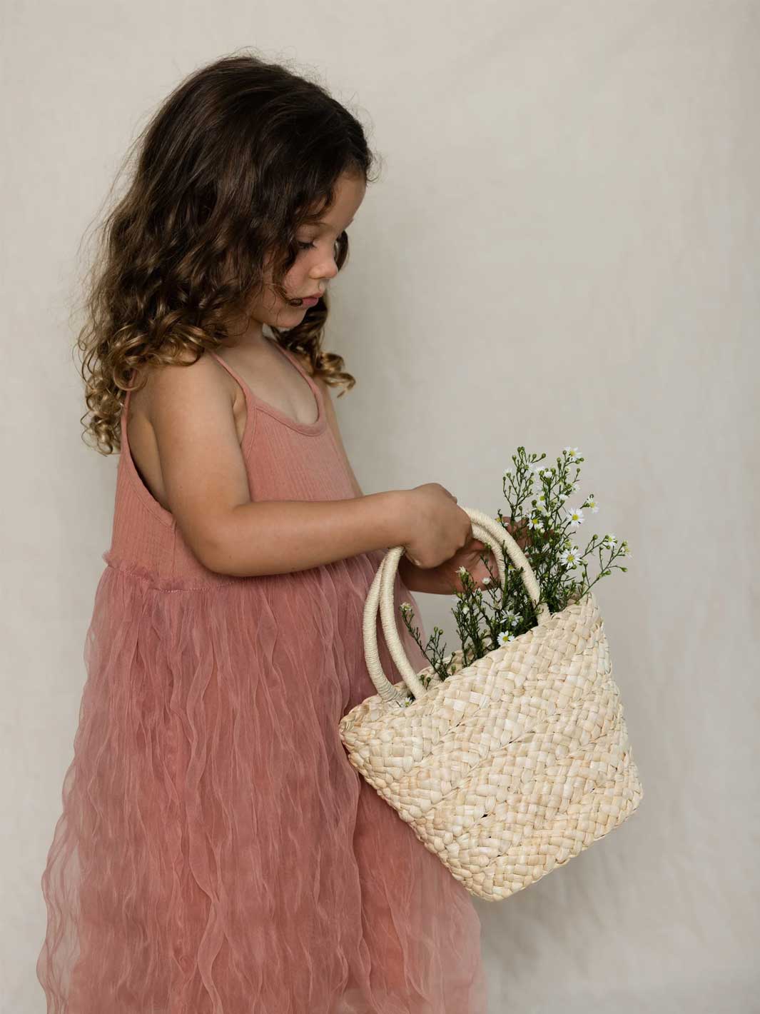 A young girl with curly brown hair wearing a dusty pink tulle dress holds a woven straw tote bag with cream handles, filled with small green and white flowers, against a neutral off-white background.