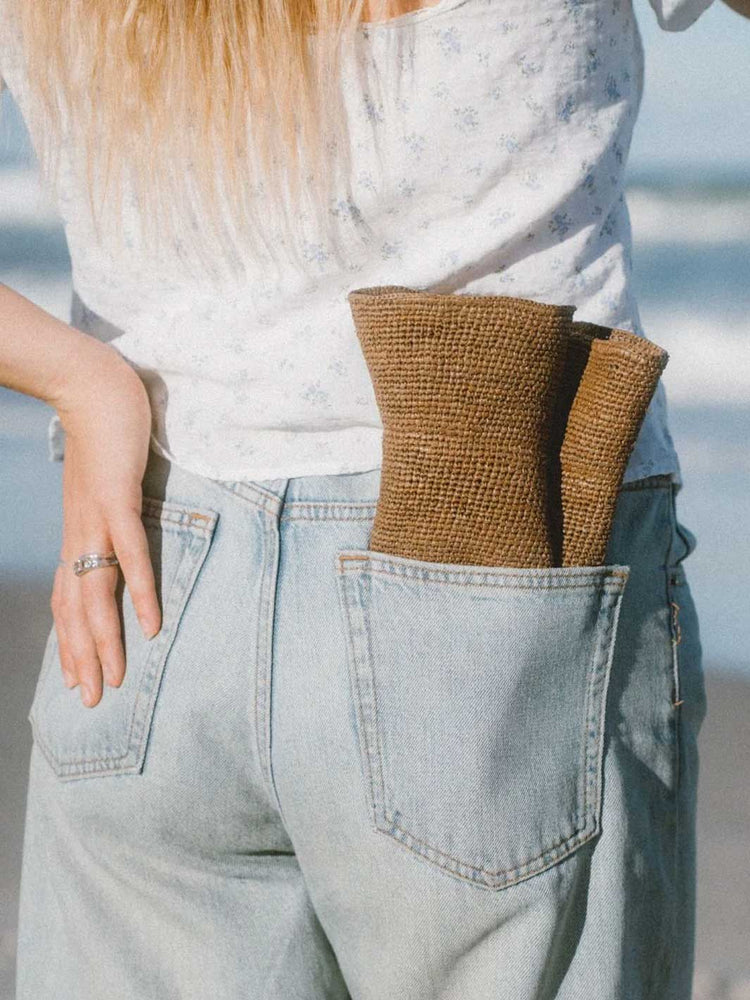 A close-up lifestyle shot of a person wearing light-wash blue jeans and a white floral top, with a brown woven burlap hat tucked into the back pocket.