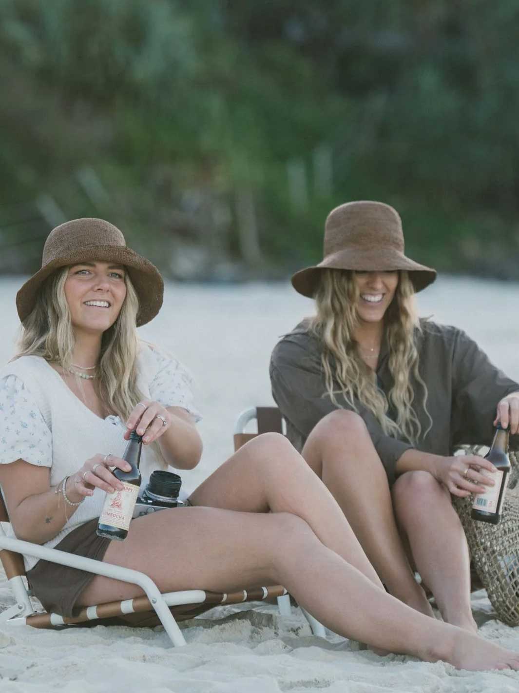 Two women sitting on beach chairs on a sandy beach, wearing wide-brimmed brown hats and holding bottles.