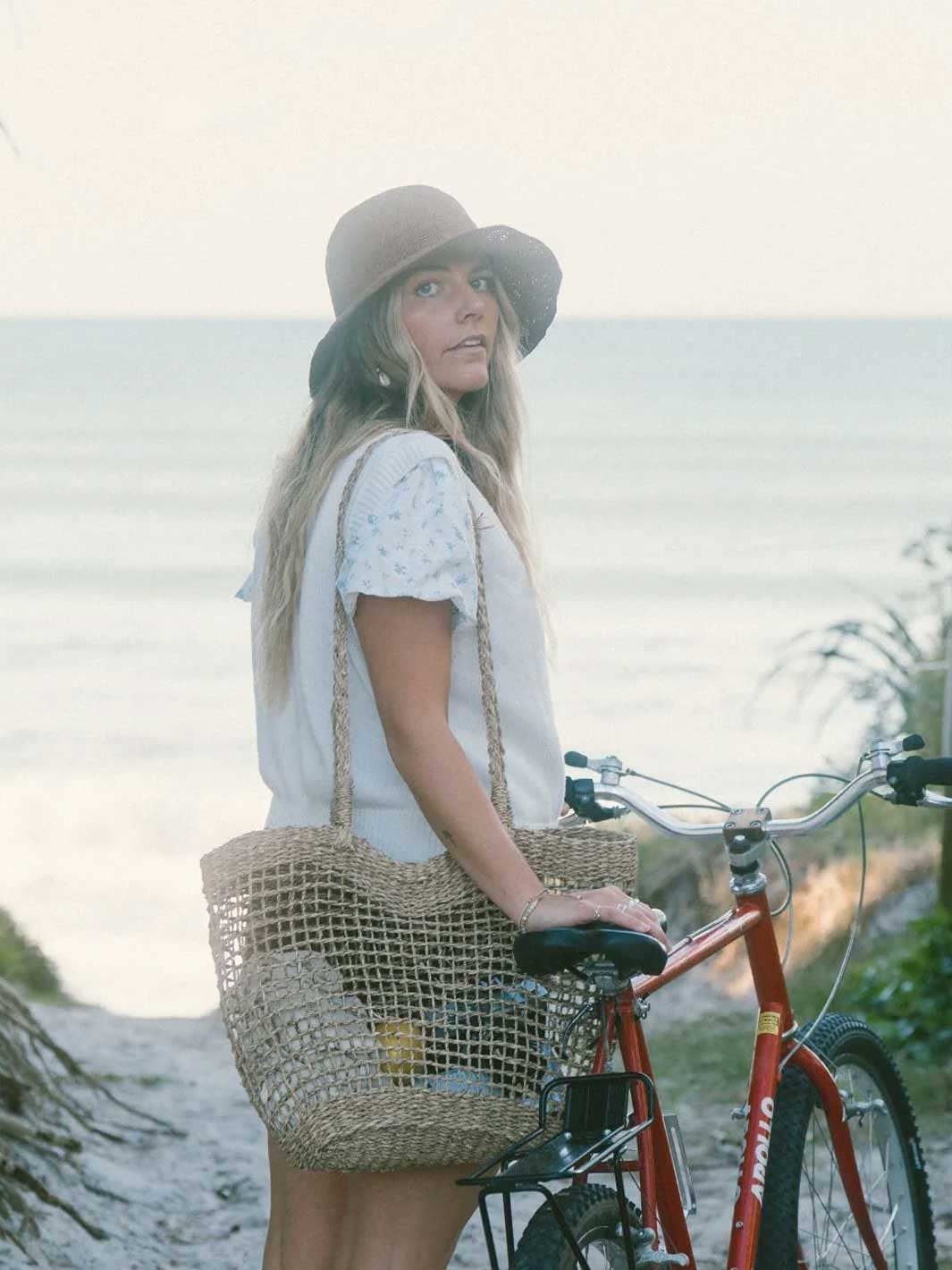 A woman wearing a wide-brimmed hat and a white top stands beside a red bicycle on a beach path, carrying a large woven straw tote bag.