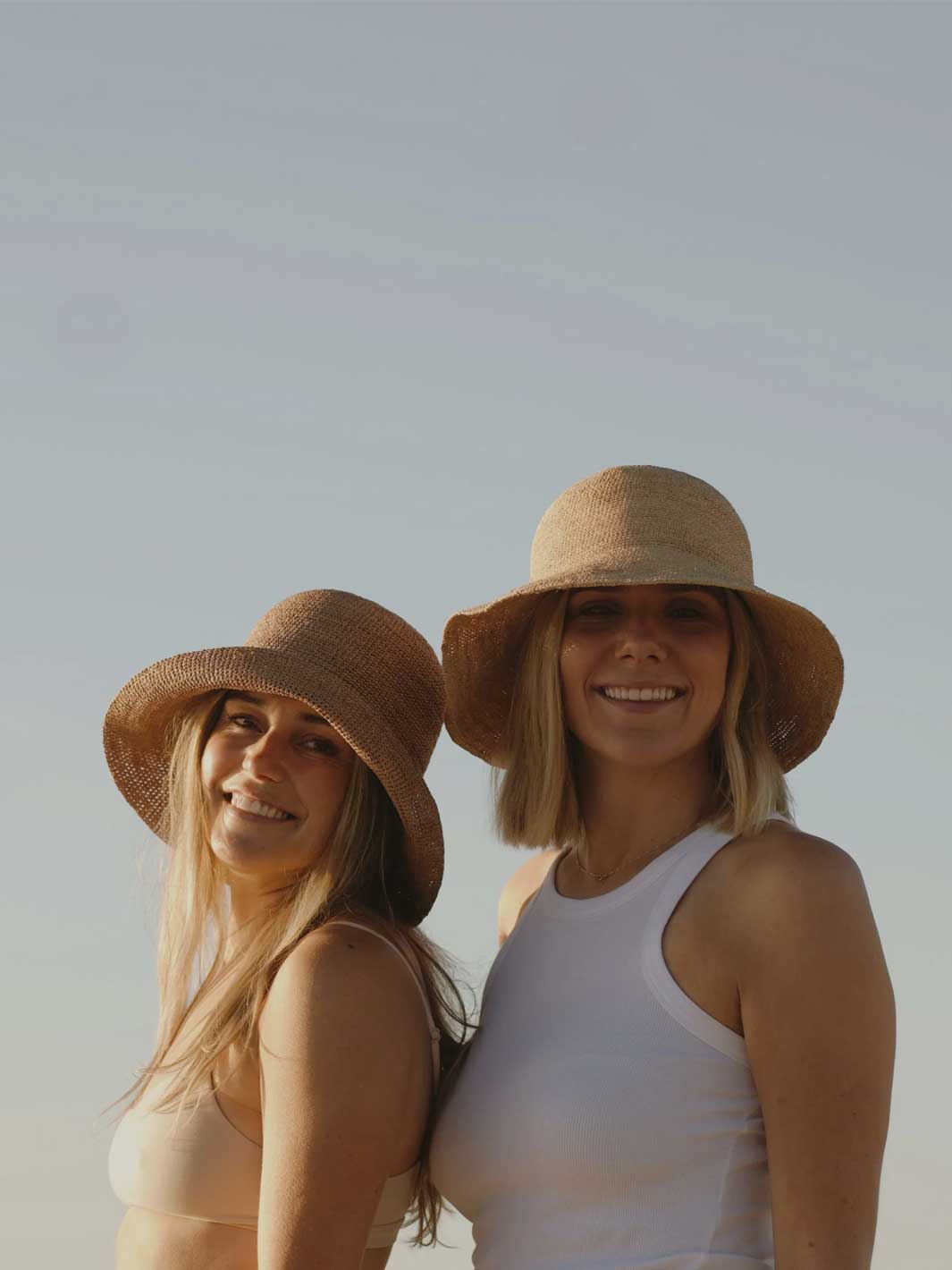Two women smiling and posing outdoors against a pale sky, each wearing a wide-brimmed straw sun hat. One wears a beige bikini top, and the other wears a white tank top.