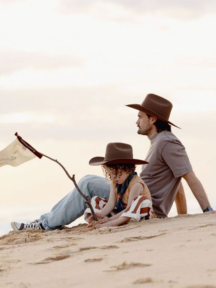 A man and a child sitting on a sandy dune outdoors, both wearing brown wide-brim cowboy hats, with the man in a gray shirt and the child in a striped top.