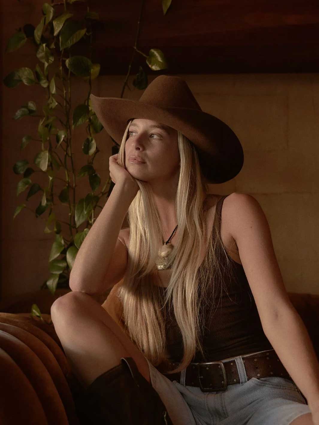 A woman with long blonde hair sits on a leather couch wearing a brown cowboy hat, a dark brown tank top, and light blue denim shorts in a warm, dimly lit room.