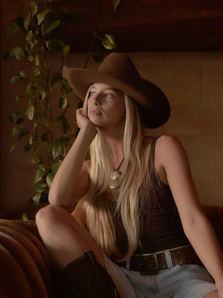A woman with long blonde hair sits on a leather couch wearing a brown cowboy hat, a dark brown tank top, and light blue denim shorts in a warm, dimly lit room.
