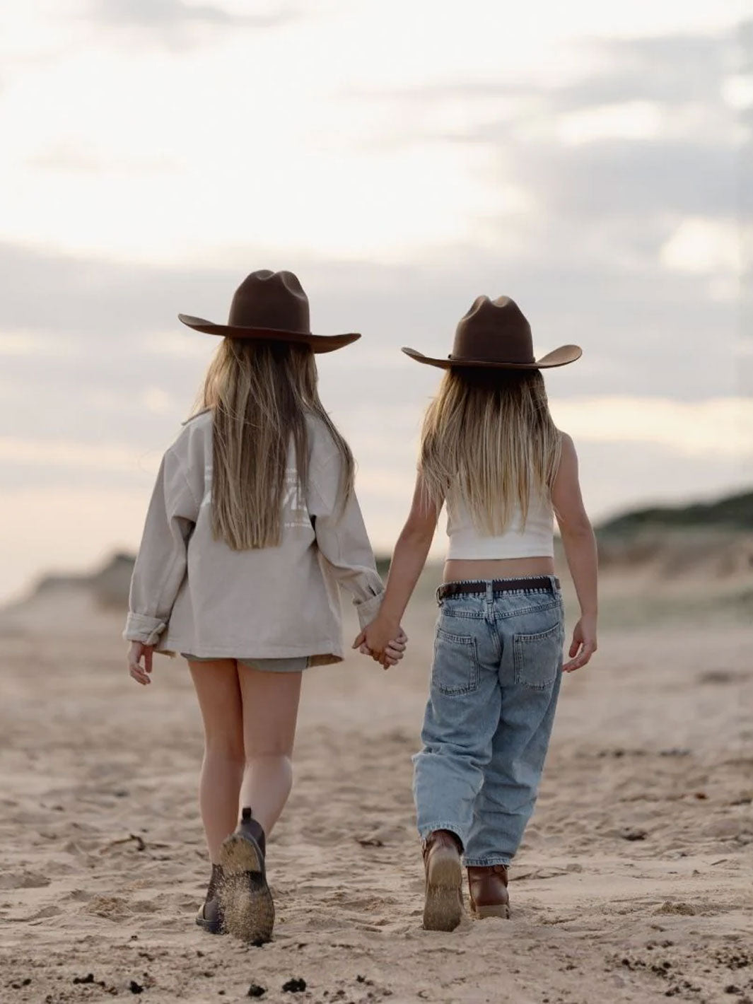 Two girls walking hand-in-hand on a sandy beach from behind, wearing brown cowboy hats, boots, a light beige shirt, and light blue jeans under a cloudy sky.