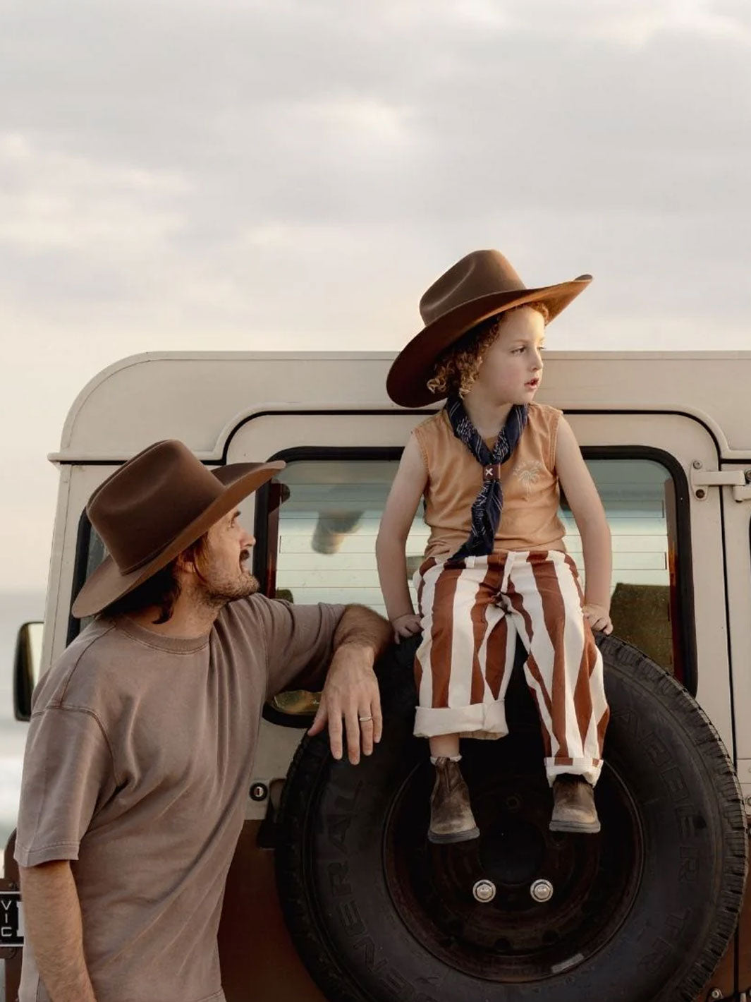 A man and a young child wearing matching brown cowboy hats and western-style clothing pose in front of a vintage off-white vehicle with a spare tire. The man wears a taupe t-shirt, and the child wears a tan sleeveless top, brown-and-white striped pants, and a dark blue neck scarf.