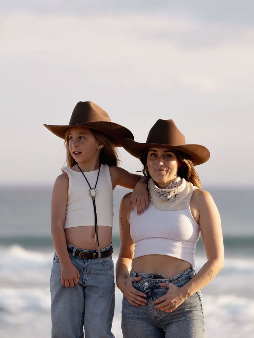 A woman and a girl standing on a beach wearing matching brown cowboy hats, white tank tops, and blue jeans, with the ocean and cloudy sky in the background.