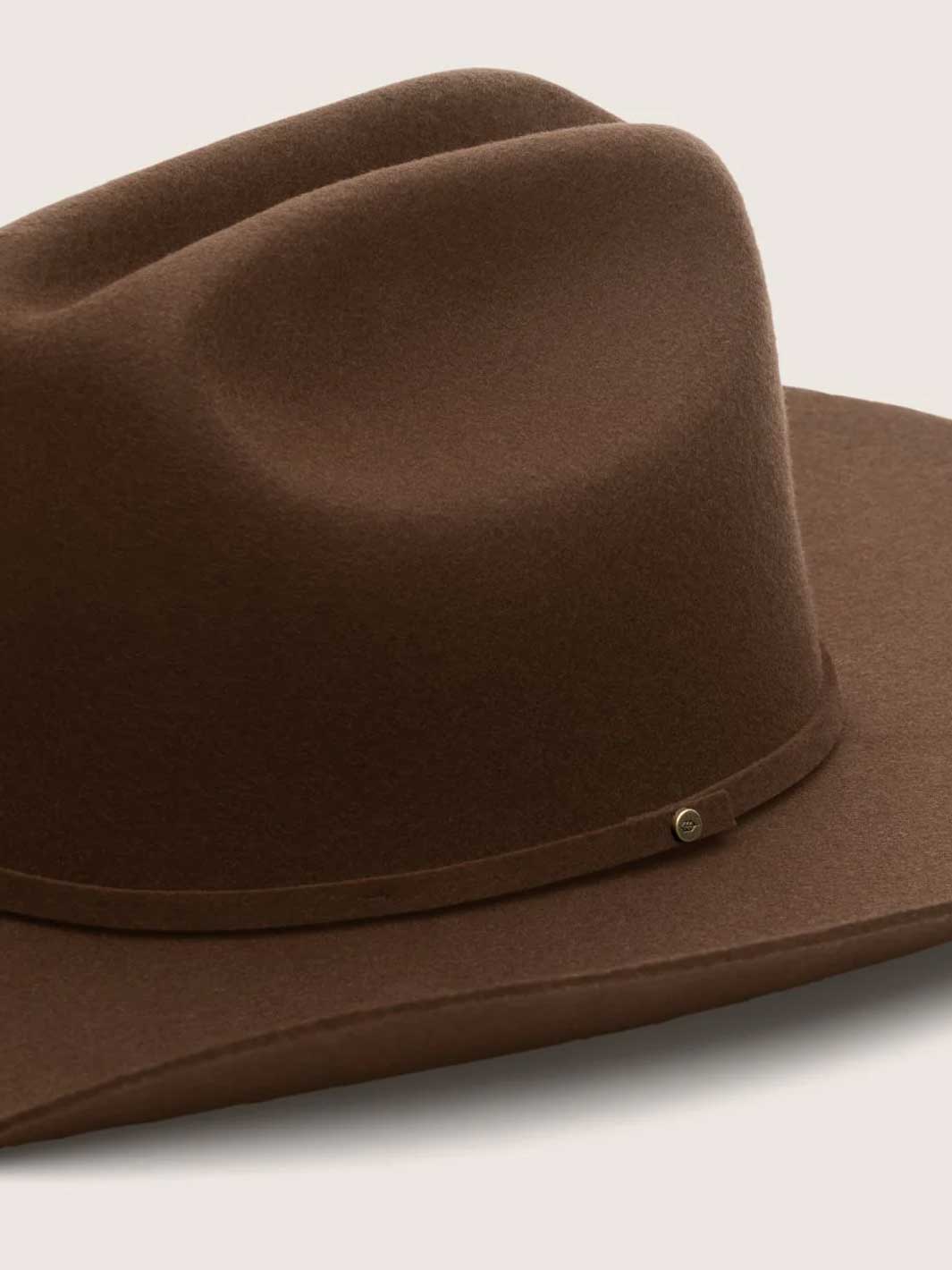A close-up of a brown felt cowboy hat with a matching band and a small metallic snap detail, set against a light beige background.