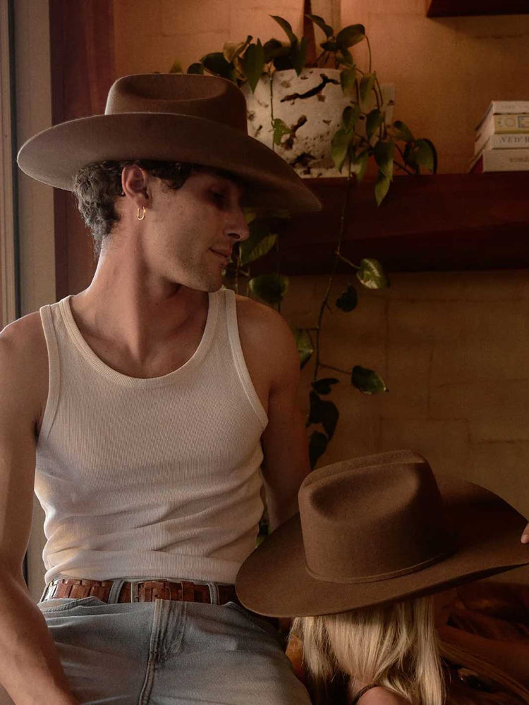 A man wearing a brown wide-brim cowboy hat and a white ribbed tank top sits indoors next to a woman wearing a matching brown cowboy hat. A potted trailing plant and a shelf with books are visible in the background.