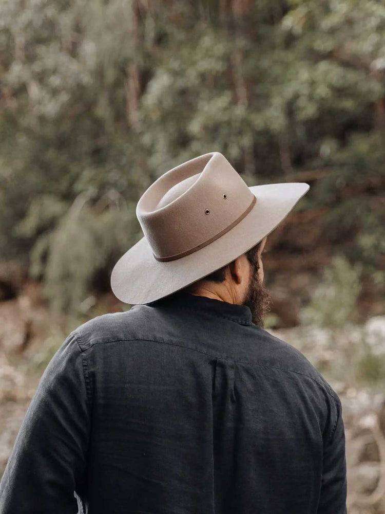 A man viewed from behind wearing a wide-brimmed taupe felt hat and a dark charcoal shirt, standing outdoors in a blurred forest setting.