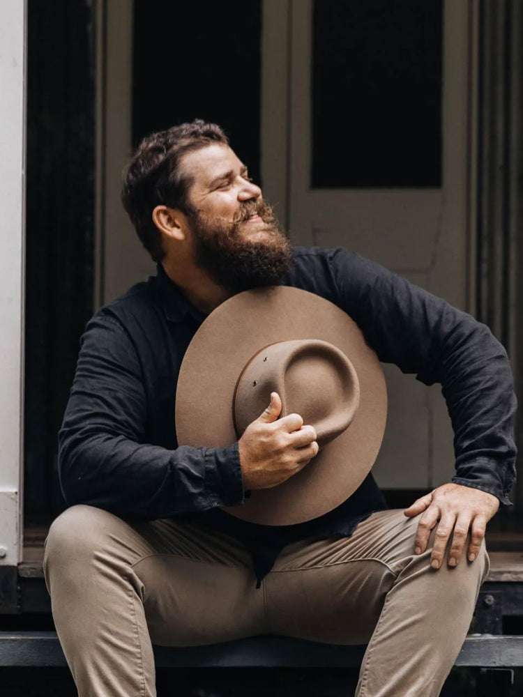 A bearded man in a black shirt and khaki pants sits on a porch step holding a large brown wide-brim hat against his chest.