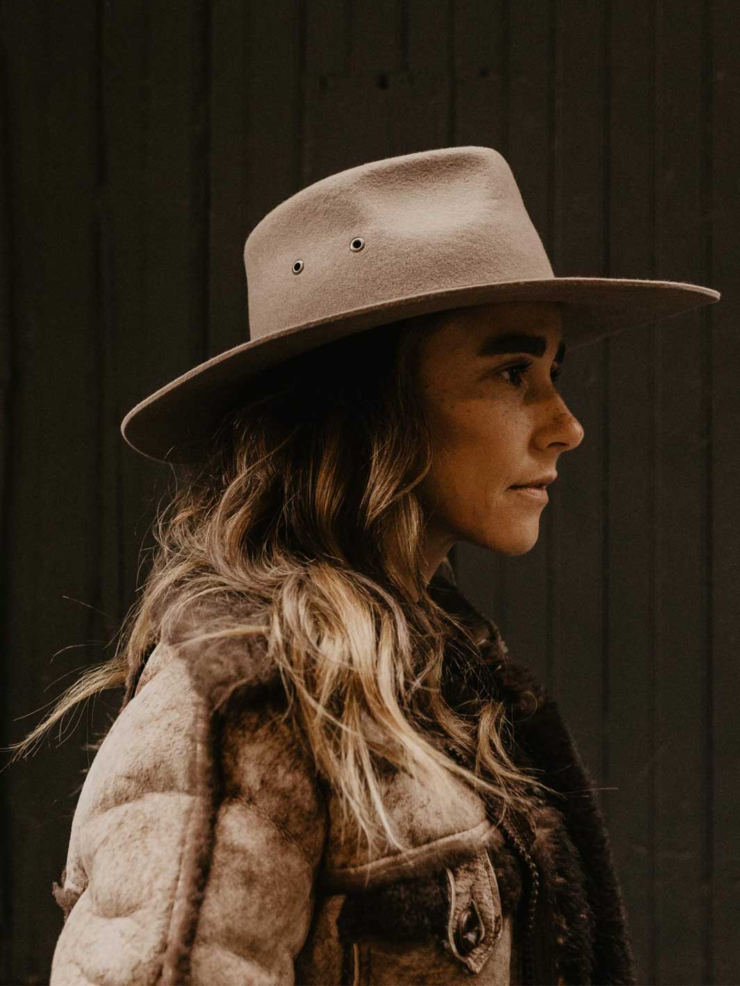 A woman in profile wearing a wide-brimmed felt hat and a textured jacket, photographed against a dark wooden plank background.