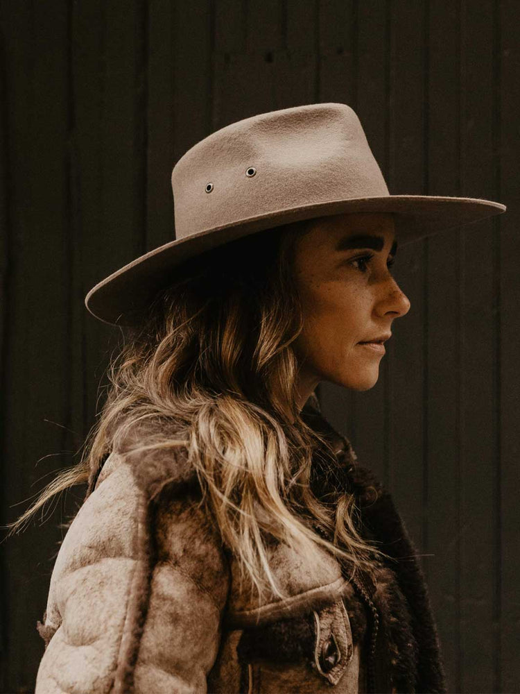 A woman in profile wearing a wide-brimmed felt hat and a textured jacket, photographed against a dark wooden plank background.