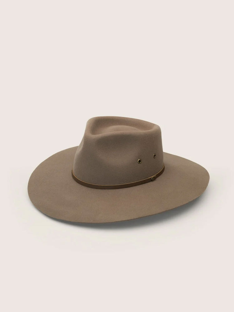 A wide-brimmed felt outback hat in a muted taupe-brown colour, featuring a dark brown band around the crown and two small metal eyelets, set against a solid light beige background.