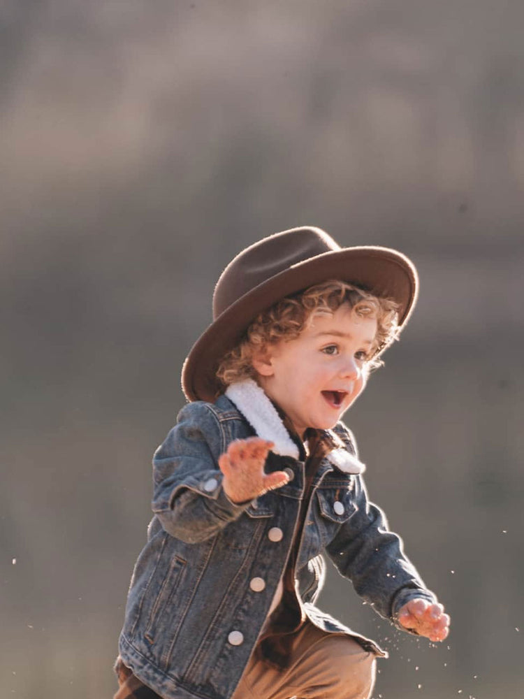A toddler wearing a brown wide-brim felt hat, a blue denim jacket with a white sherpa collar, and tan pants, captured mid-motion against a soft, blurred outdoor background.