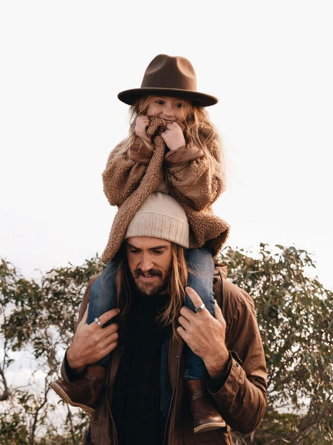 A man wearing a brown jacket and a cream beanie carries a young girl on his shoulders. The girl is wearing a brown felt fedora hat and a brown teddy fleece jacket.