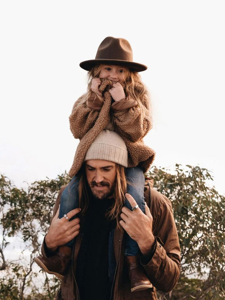 A man wearing a brown jacket and a cream beanie carries a young girl on his shoulders. The girl is wearing a brown felt fedora hat and a brown teddy fleece jacket.
